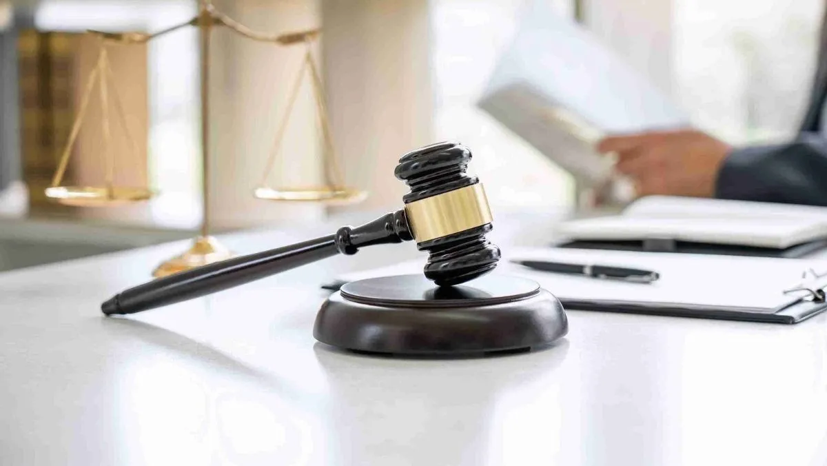 A gavel resting on a desk, with a backdrop featuring a man reading a book and a justice scales