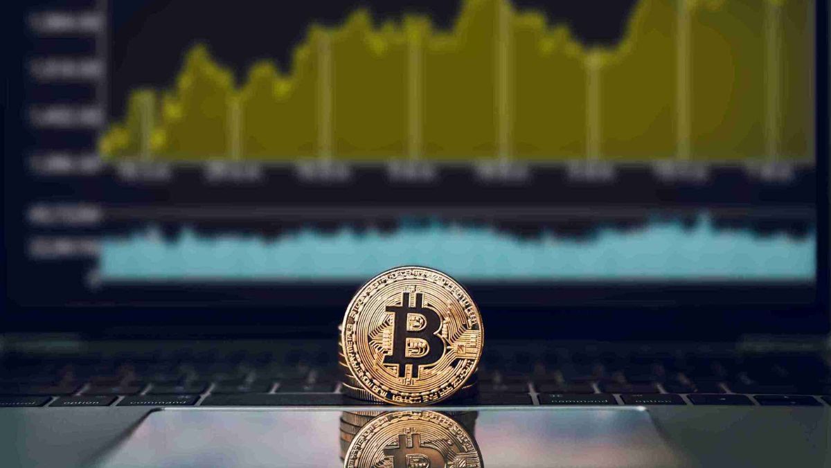 A golden Bitcoin placed on the trackpad of a laptop with a screen displaying a trading chart