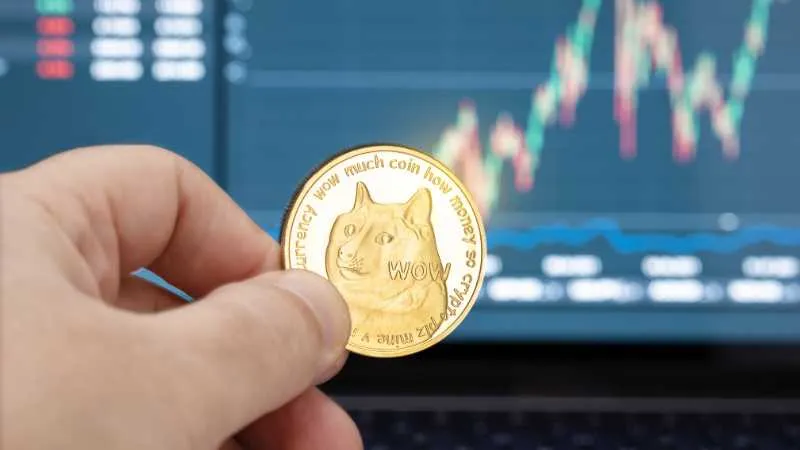 A hand holds a Dogecoin in front of a computer screen with trading charts.