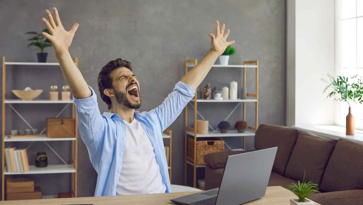A happy guy cheers with his hands up looking at the laptop on the table