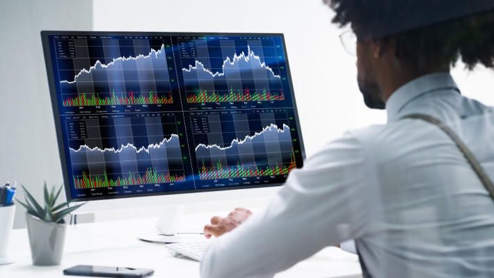 A male trader sits in a chair, observing trading charts on computer screens