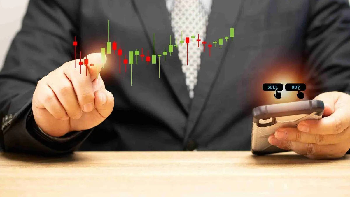A man gestures towards a candlestick pattern, holding a mobile phone featuring buy and sell options