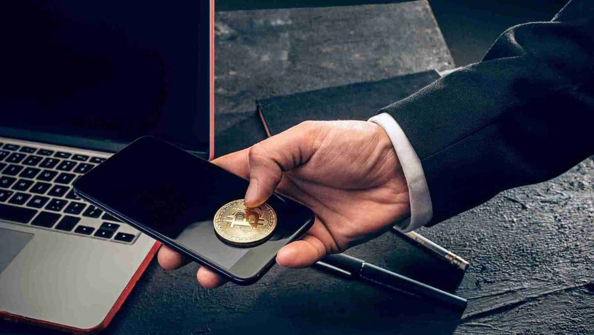 A man holds a mobile phone featuring a gold Bitcoin token with a laptop and pen placed on the table