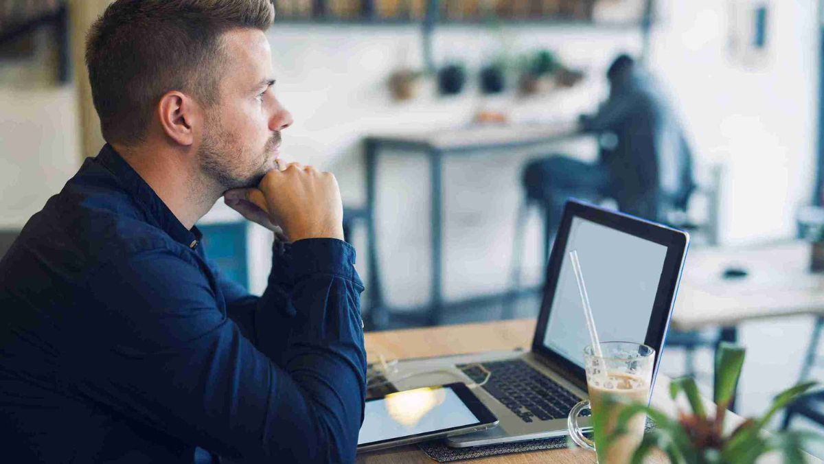 A man in a cafe, seated in front of his laptop, deep in thought