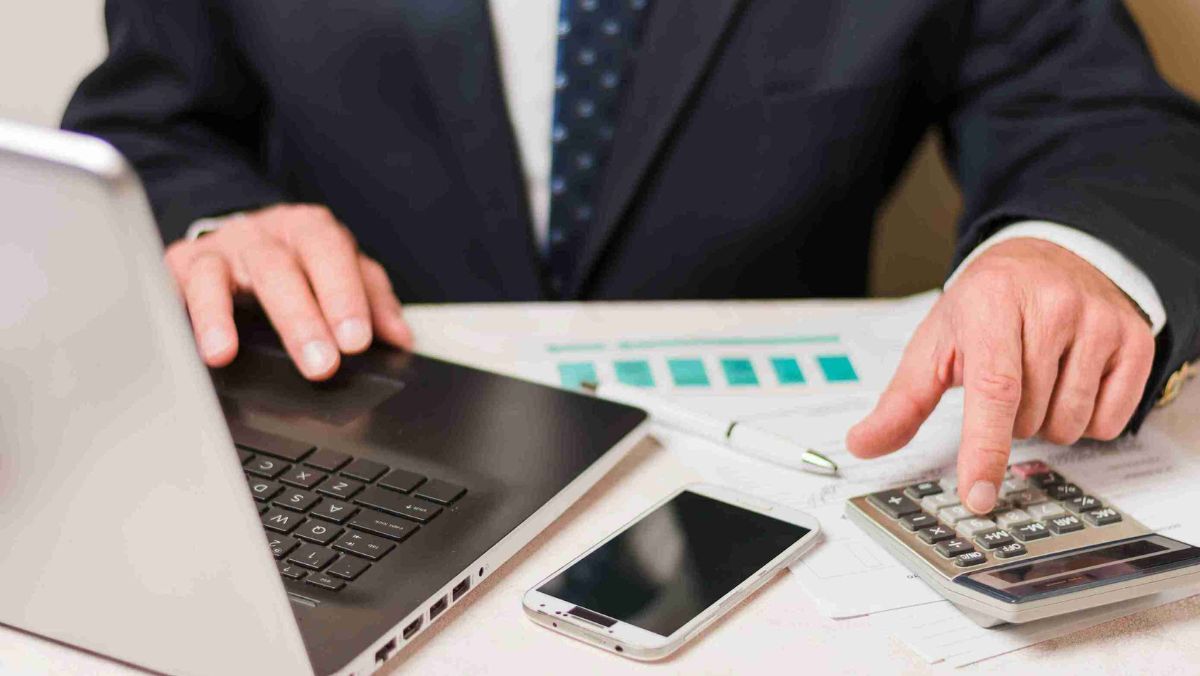 A man in a suit working on his desk with a laptop, calculator, mobile phone, and paper documents