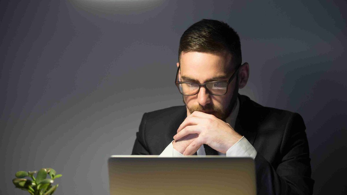 A man in eyeglasses thinking deeply in front of his laptop with a low light background