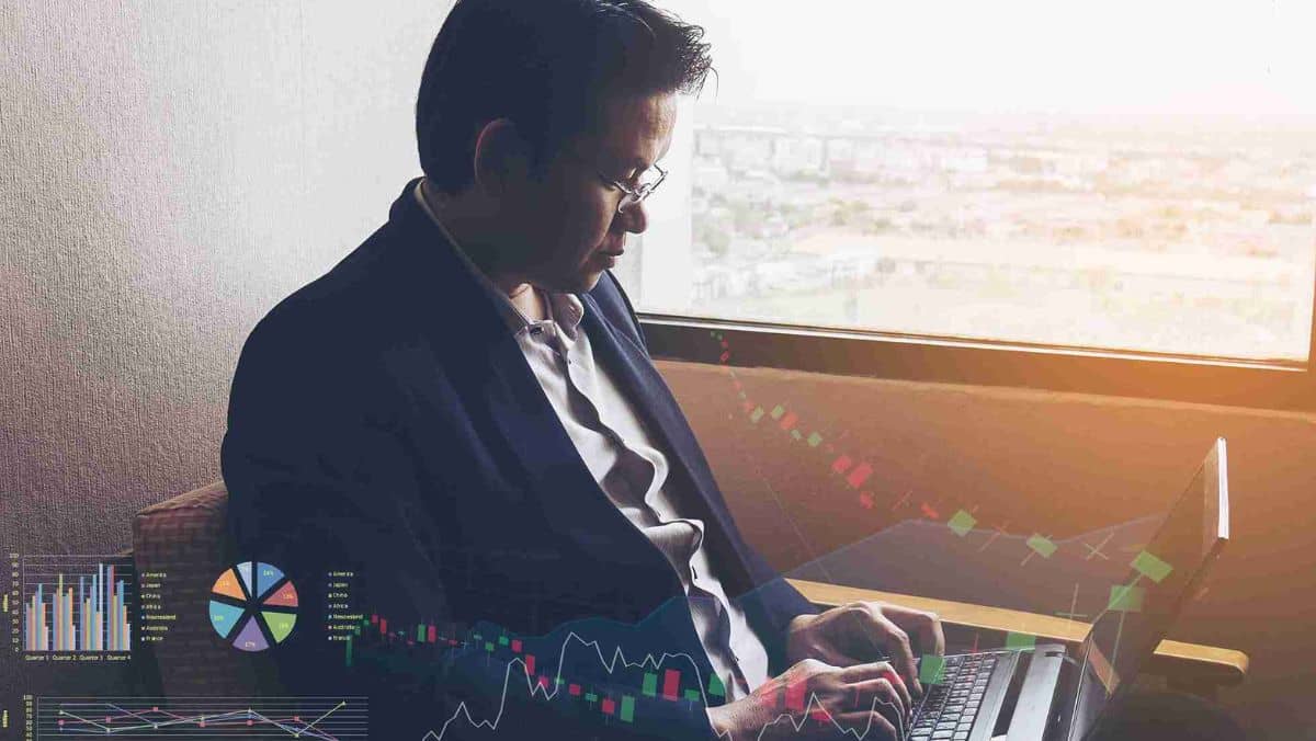 A man in formal attire carefully analyzing information on his laptop with trading patterns visible in the background