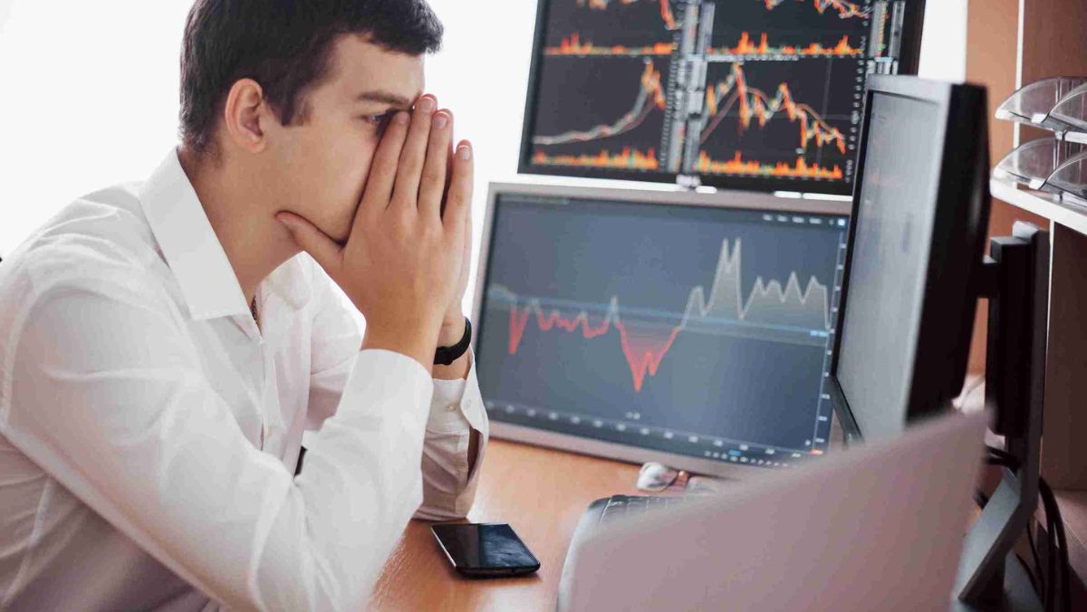 A man in white clothes putting his hands on his face showing frustration in front of multiple computer screens 