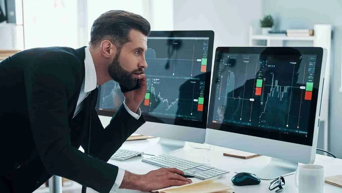 A man talks on his phone while simultaneously monitoring multiple trading charts on computer screens