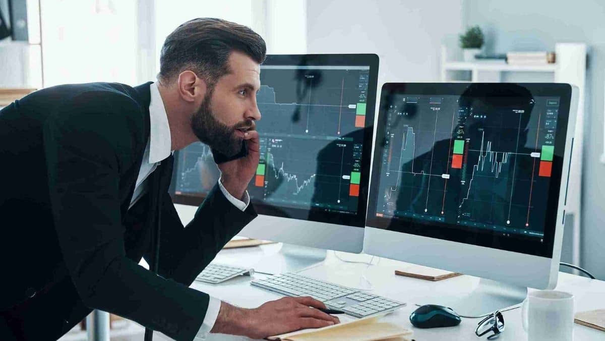 A man talks on his phone while simultaneously monitoring multiple trading charts on computer screens