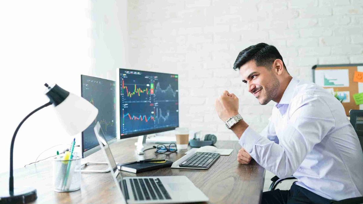 A man working in his office rejoicing in front of multiple computer screens and a laptop