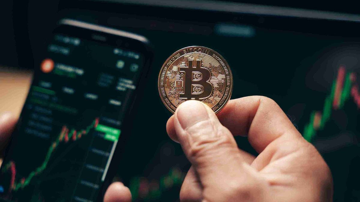 A mobile device held for trading in one hand, and in the opposite hand a single Bitcoin being held up