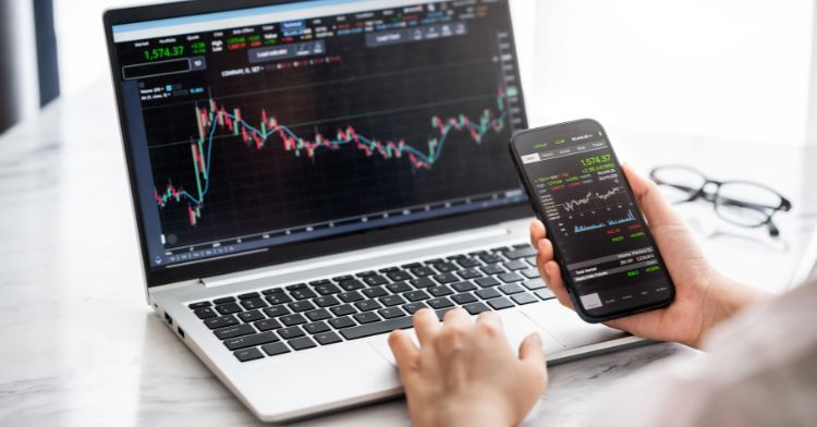 A person analyzing financial data on a laptop screen with stock market graphs, while holding a smartphone displaying a trading app.