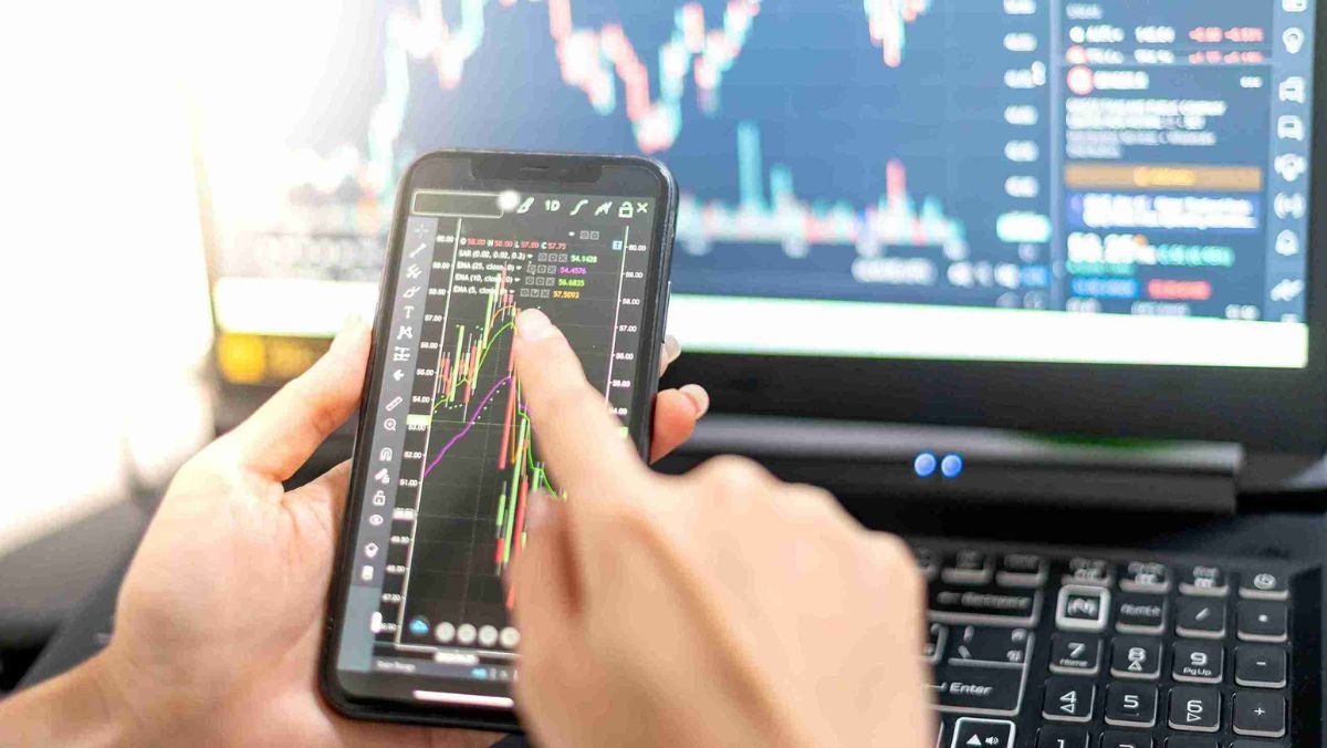 A person directs attention to a live trade on a mobile device while a computer on the table presents financial charts