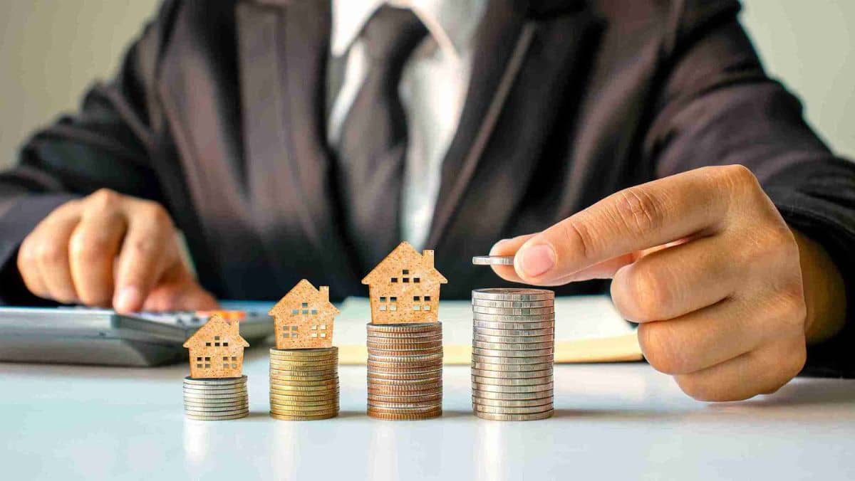 A person in a black suit stacking coins with a wooden flatshaped house