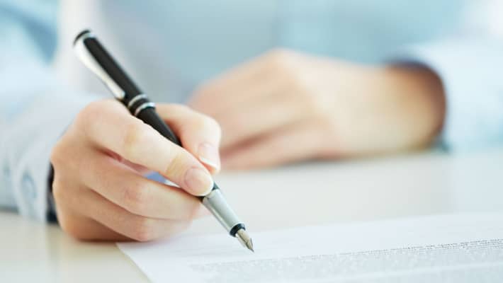 A person in a blue sweater signing a document on a clipboard with a silver pen