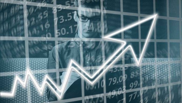 A person situated centrally, jotting down notes with an upward arrow, surrounded by stock market data in the background