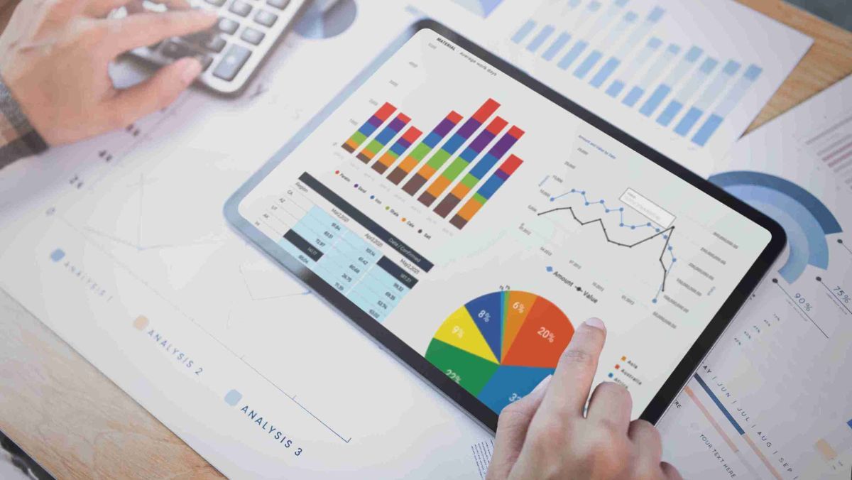 A person using a tablet to show financial data surrounded by documents and a calculator placed on the table