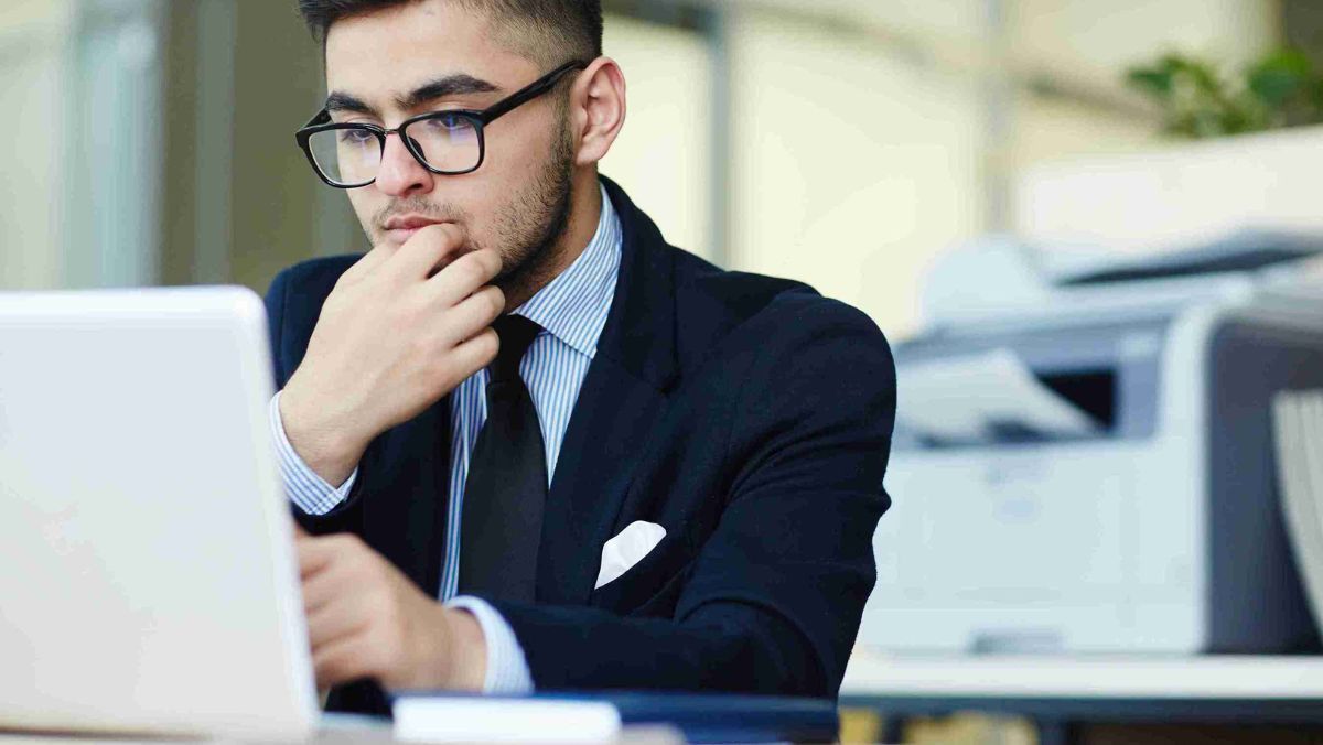 A person wearing a business suit and eyeglasses immersed in thought while observing the laptop screen