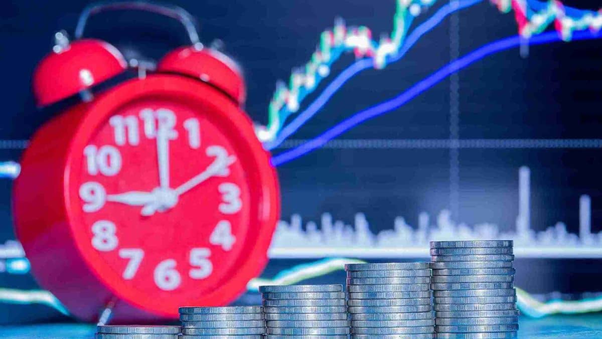 A red alarm clock and a stack of coins positioned against a trading chart