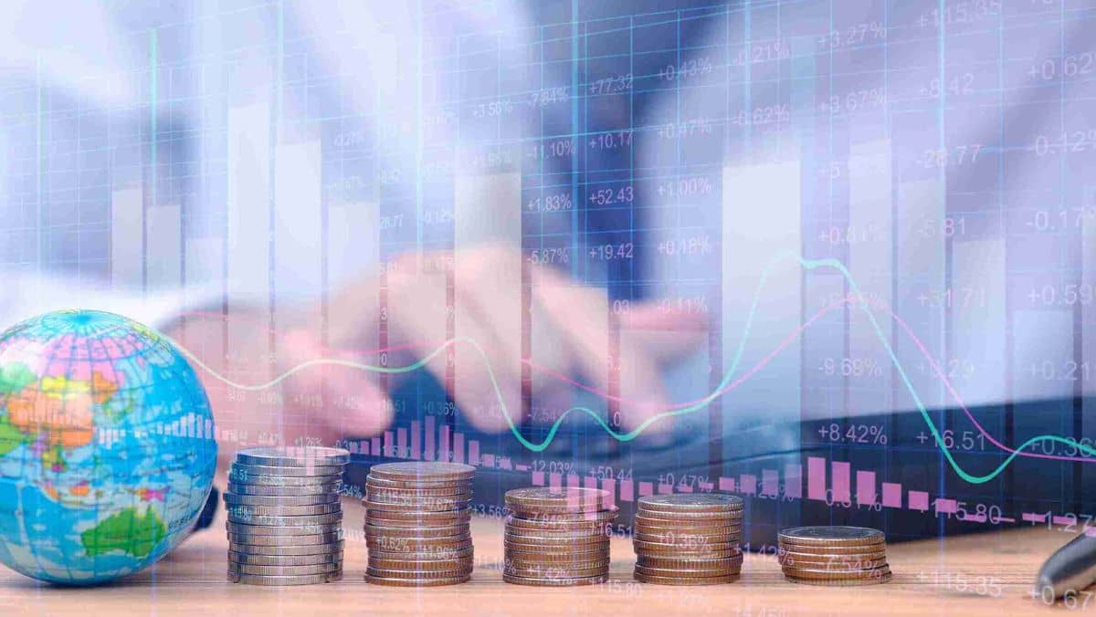 A table with a stack of coins a globe and a trading chart in the backdrop