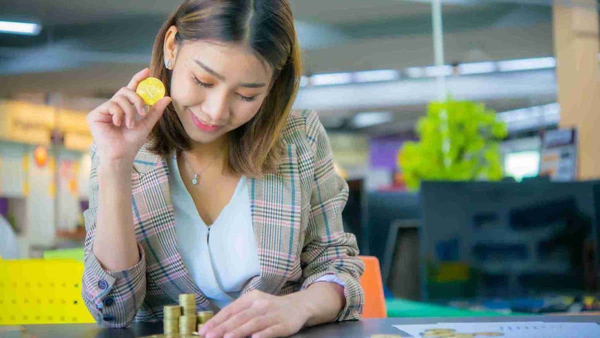 A woman is smiling while looking at a table showcasing a stack of gold coins, her hand holds one piece of gold coin