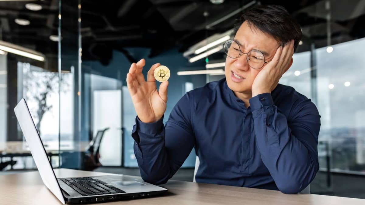 Confused individual holding a Bitcoin, contemplating it while seated in an office setting with a laptop