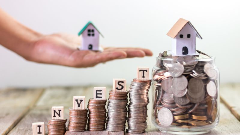 Hand hovering over coins stacked into the word 'INVEST', with a house model on top and in a jar, representing real estate investment