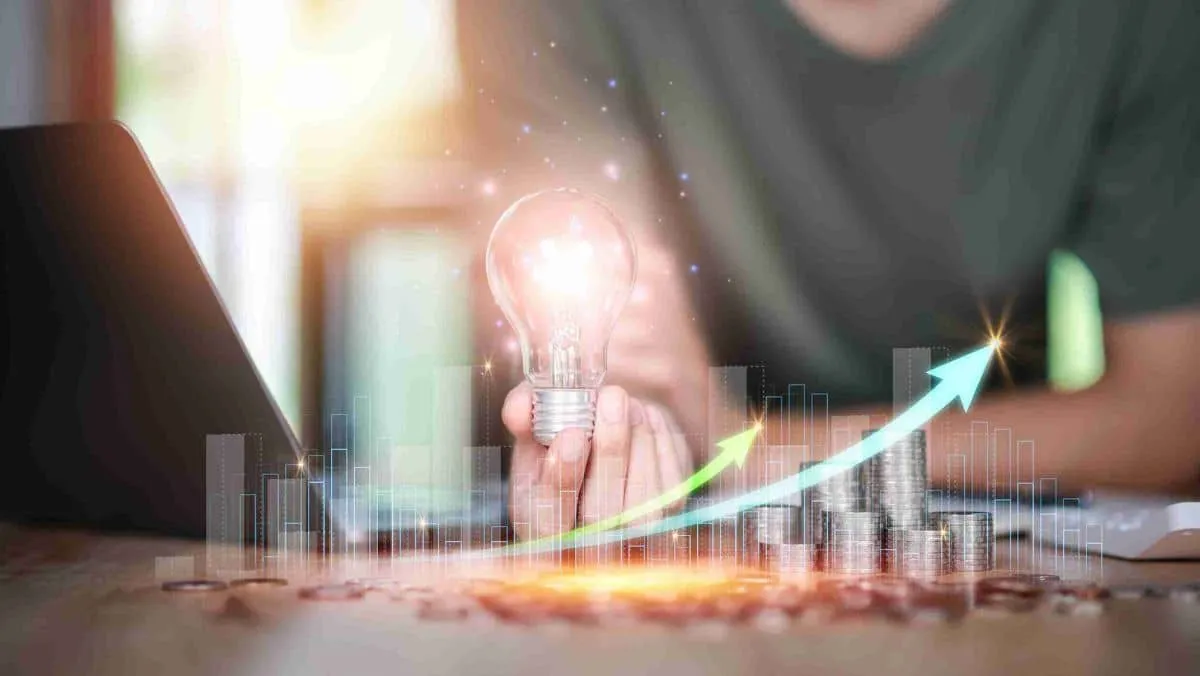 Man holding a light bulb while surrounded by coins on the tabletop, a laptop, and a visual representation of bar graphs