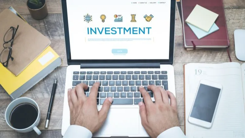 Person using a laptop with 'INVESTMENT' displayed on the screen, surrounded by office supplies.