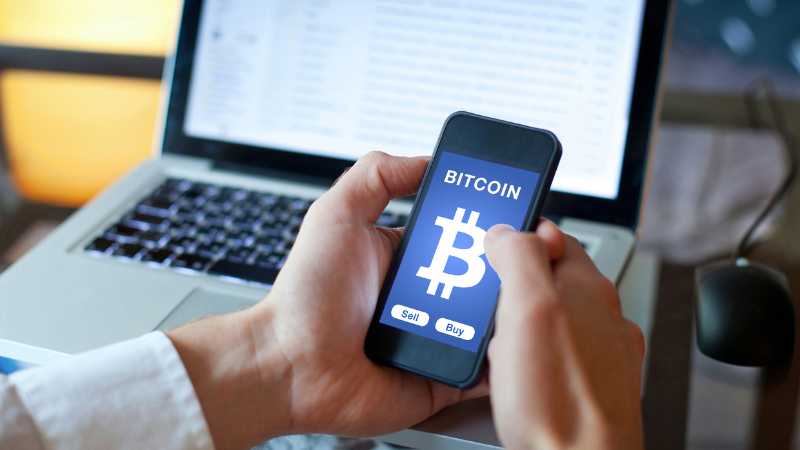 Person using a smartphone with a Bitcoin trading app in front of a laptop