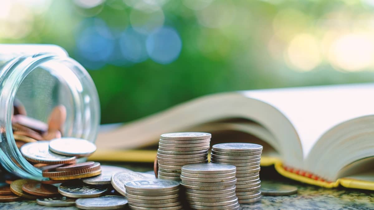 Scattered and stacked coins and a glass jar with an open book on the side