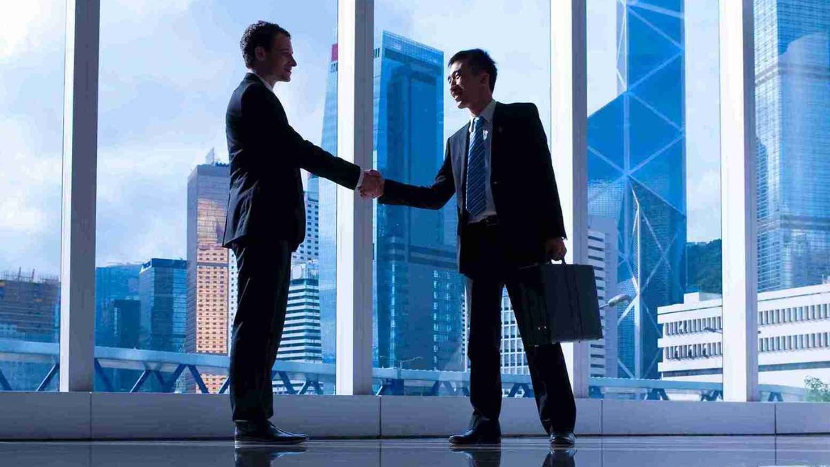 Two individuals shaking hands in an open area, with a glass wall in the background revealing buildings outside